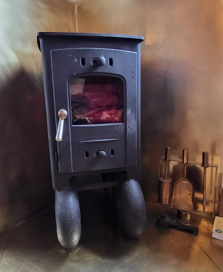 A black wood-burning stove with a small window displaying red flames is positioned in a corner, enclosed by a brass-colored metallic wall. On the right side, there is a set of fireplace tools, including a shovel, brush, and poker—giving the room the cozy charm of The Quarterdeck.