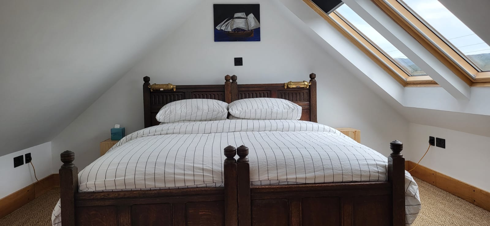 A cozy bedroom in The Quarterdeck features a wooden double bed placed against an angled white wall. The bed has striped bedding and a dark wooden headboard. Above the bed is a boat artwork. A skylight window on the right lets in natural light, and two bedside tables with lamps complete the setting.