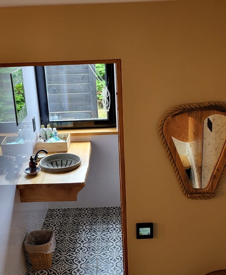 A room at The Quarterdeck with a view of a bathroom featuring a ceramic sink on a wooden countertop. The floor has patterned tiles, and a wicker wastebasket is placed below the sink. A heart-shaped mirror is mounted on the beige wall near a wooden chair.