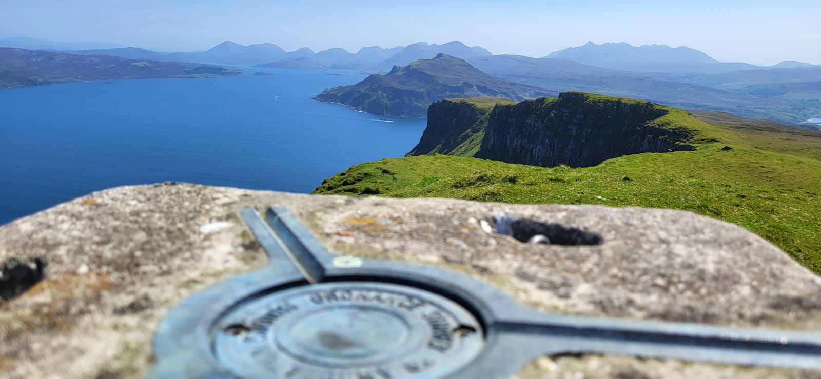 From the mountain summit, a circular trig point stands at the forefront like a quarterdeck. Vast blue ocean waters meet rolling green hills and rugged cliffs in the midground. Distant mountains fade into the hazy sky, forming a serene panorama.