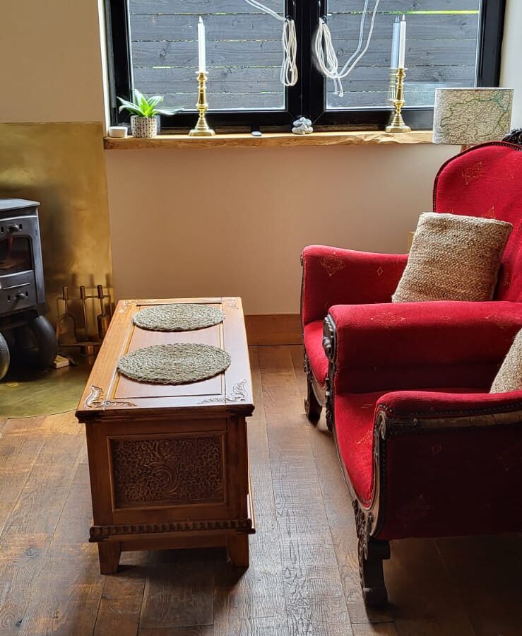 A cozy living room with a red armchair and a wooden coffee table on a hardwood floor. The armchair has a cushion, and the coffee table holds a tray. Two tall candles stand on the windowsill, along with decorative items reminiscent of The Quarterdeck. Sunlight filters through the window.