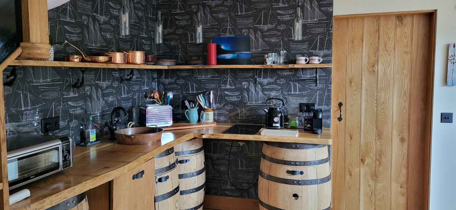 A cozy kitchen with a wooden theme, aptly named The Quarterdeck, features a countertop made of repurposed barrels. The walls have nautical-themed wallpaper. The counter holds various items including a toaster, kettle, coffee machine, cups, and cooking utensils. A wooden door is on the right.