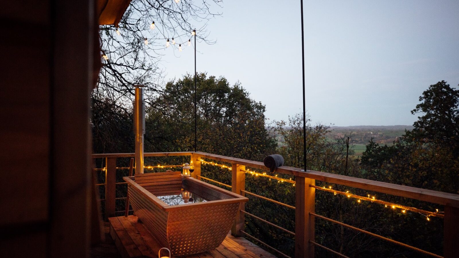 A cozy outdoor wooden deck at Pennard Hill is decorated with string lights and features a hot tub. The deck offers a scenic view of tree tents nestled in a lush, green landscape at dusk. A lantern is placed on the deck floor, adding to the warm ambiance.