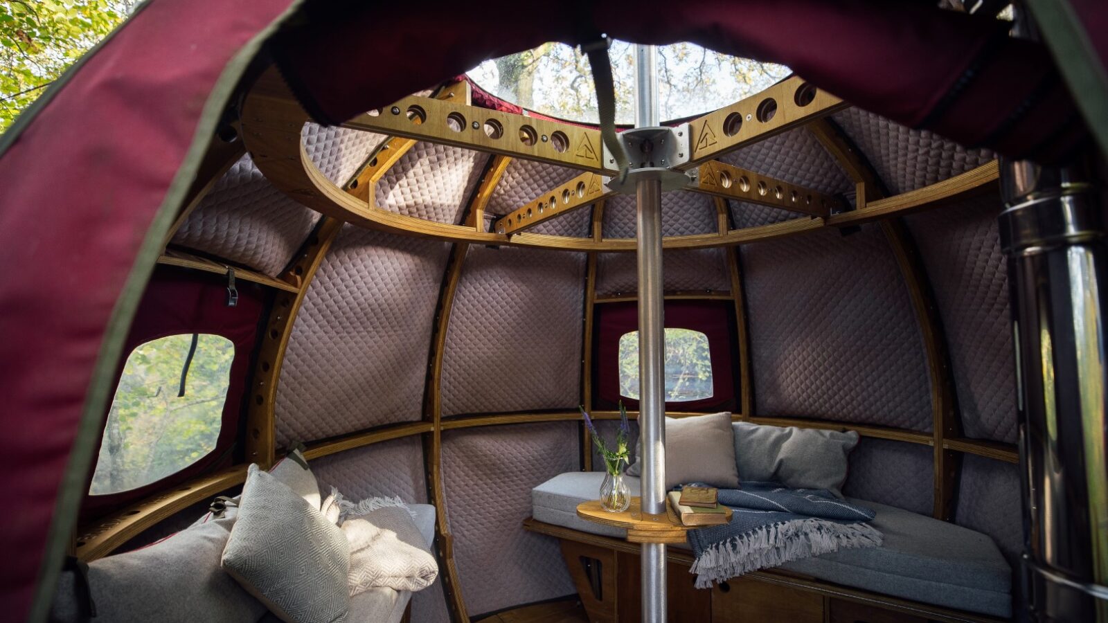 The interior of a cozy, round glamping tent at Pennard Hill boasts a wooden frame and soft, cushioned walls. It features a central pole, windows with outside views, a comfortable seating area with cushions and blankets, and a table adorned with a vase of flowers and a book.