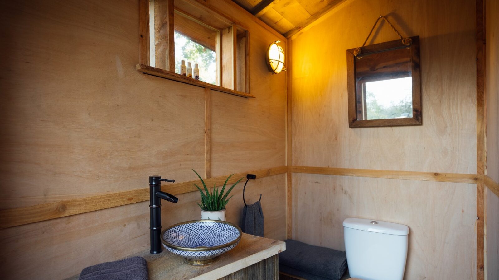 A small, rustic bathroom reminiscent of Pennard Hill with wooden walls features a white toilet, a wooden counter with a patterned basin sink, and a black faucet. A mirror with a wooden frame hangs above the sink. A towel and potted plant are on the counter, while a small window provides natural light.