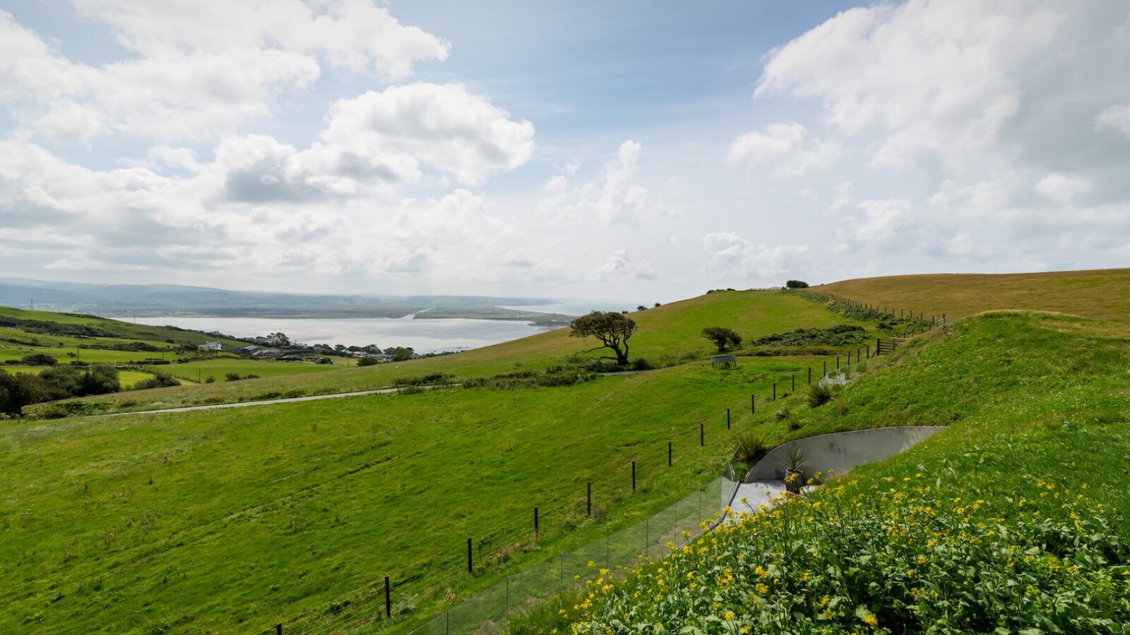 A scenic landscape features rolling green hills, a winding road, and a large body of water in the distance under a partly cloudy sky. A lush field with yellow flowers is in the foreground, separated by a fence near Willow Hill Hotel. Sparse trees dot the terrain.