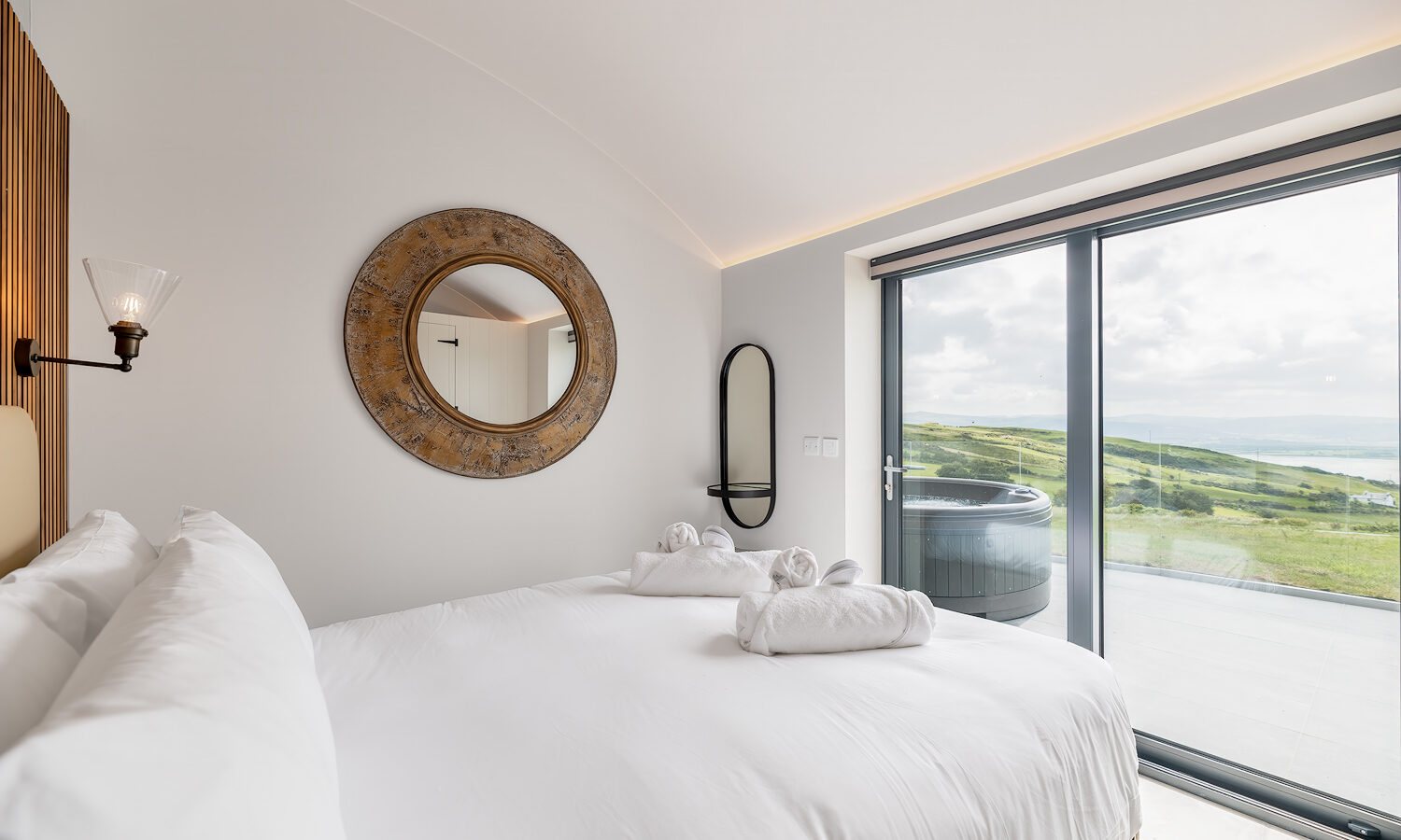 Experience a luxury stay in this modern bedroom at Willow Hill, featuring a large window with scenic views of rolling hills and a shimmering body of water. The room offers white-linen bedding, a circular mirror, wall-mounted lamp, and a balcony with a hot tub visible through the sliding glass door.