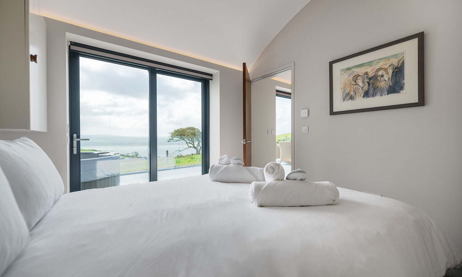 A bright bedroom with a large bed covered in white linens and two folded towels. Glass doors lead to a balcony with a scenic view of Willow Hill's green landscape and distant water. Walls are light-colored, with a painting hanging on one side and a mirror nearby.