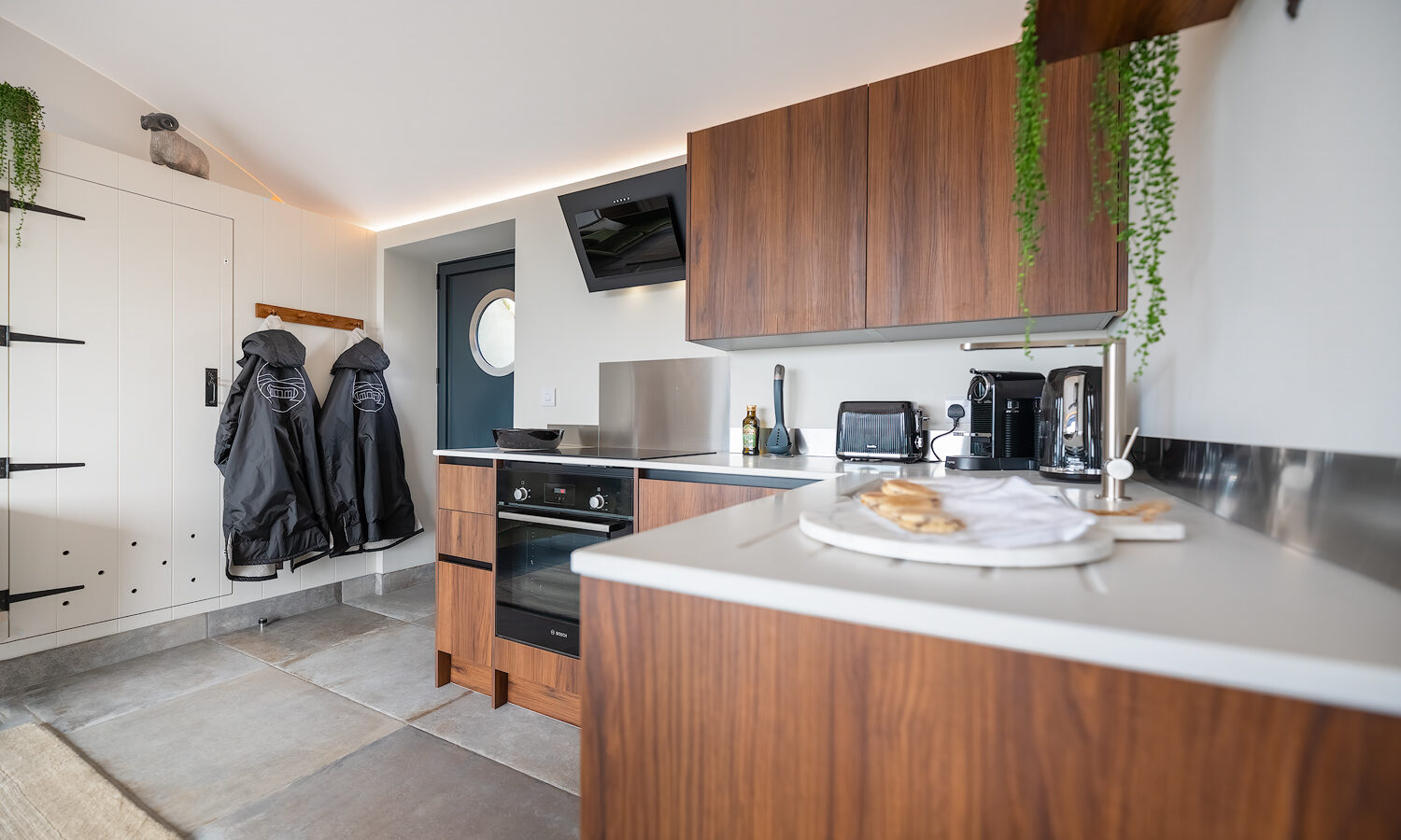 A modern kitchen at Willow Hill features wooden cabinetry, stainless steel appliances, and ceramic tile flooring. The countertop holds a coffee maker, toaster, and a plate with food. Coats hang on hooks by the door, while potted plants add greenery to the space.