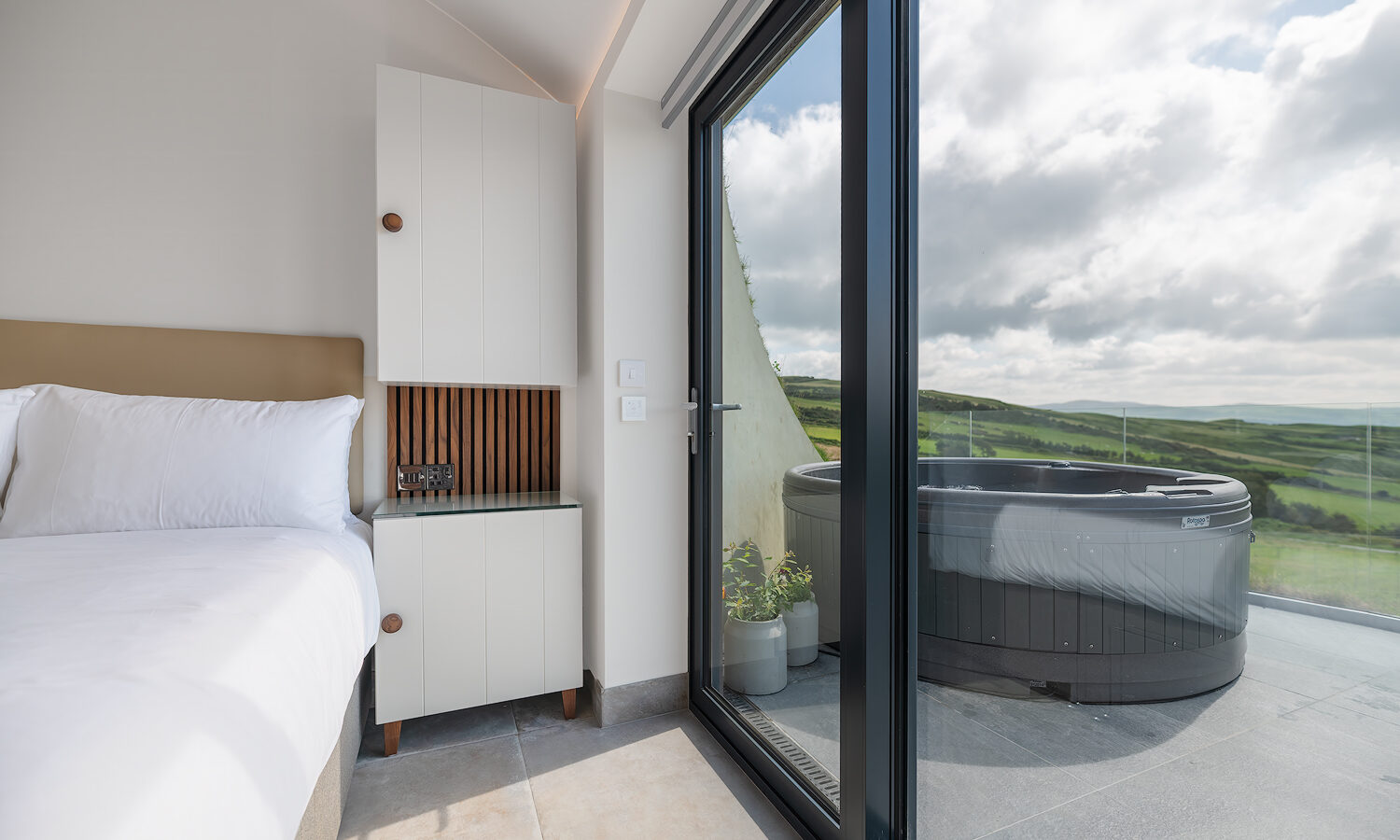 A modern bedroom at The Willow Hill features a glass door leading to a balcony with a hot tub overlooking a lush, green landscape. The room includes a white bed, a bedside table with drawers, and a small shelf. Natural light streams in brightly from the glass door, creating an inviting ambiance.