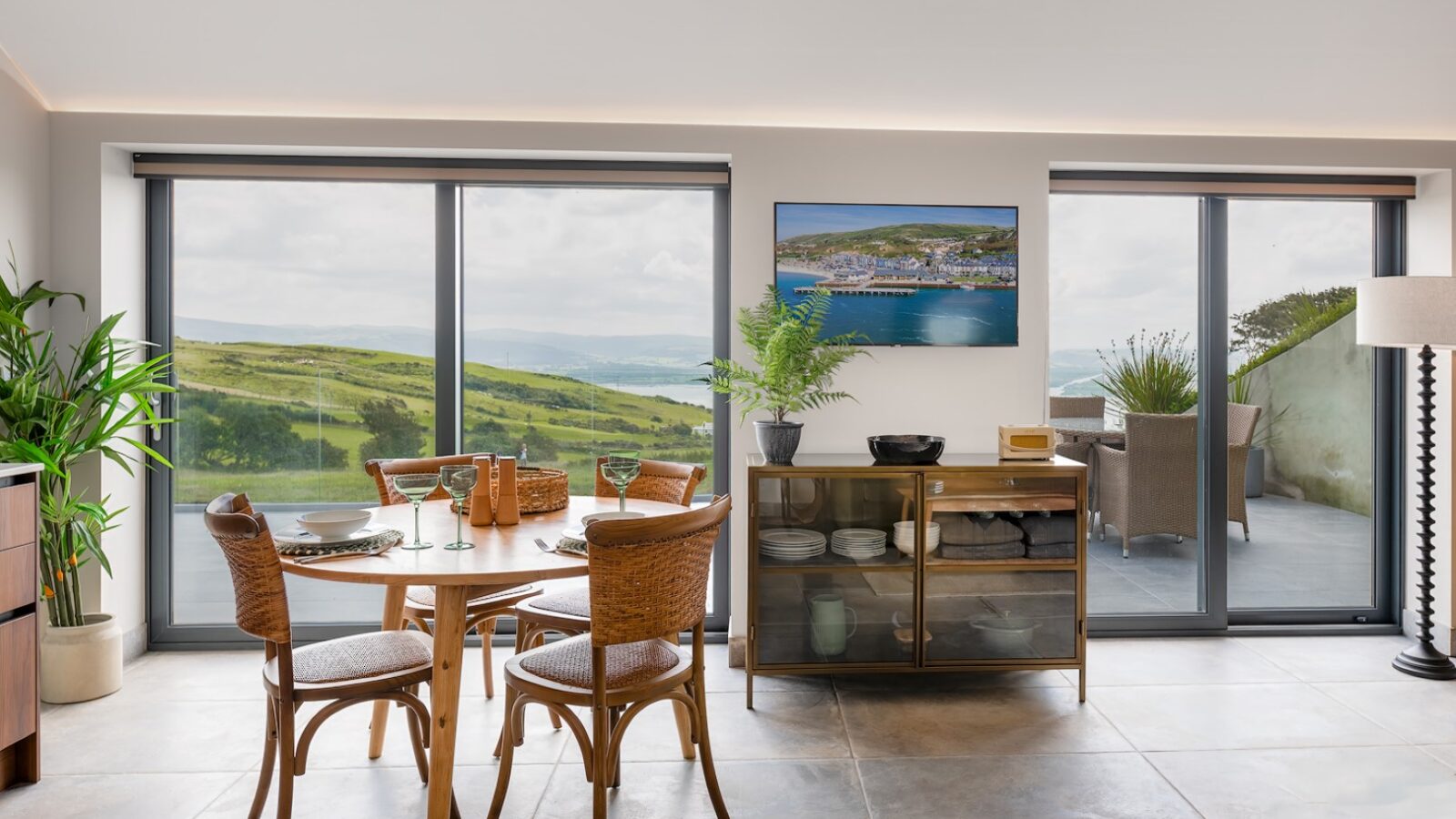 A modern dining area with a round wooden table set for four, surrounded by wicker chairs. Large windows and sliding glass doors offer a scenic view of The Willow Hill's rolling hills and a distant body of water. A cabinet with plants and decorative items sits against the wall.