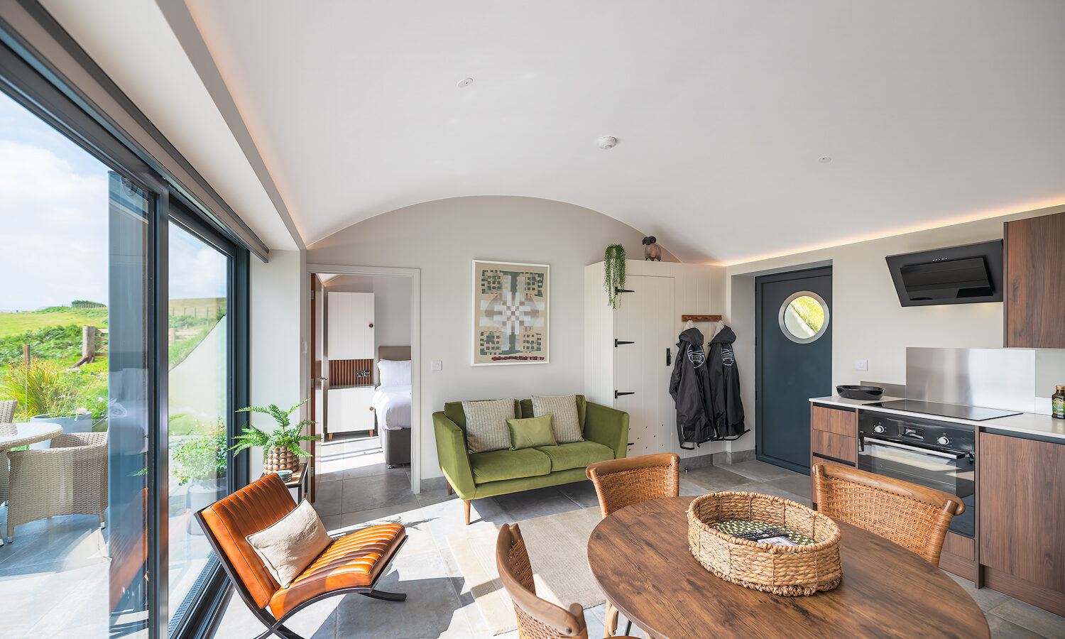 A modern, sunlit kitchen and living area at The Willow Hill features tall glass sliding doors on the left, a wooden dining table and chairs, a green couch against the back wall, and a kitchen area with an oven. Wooden accents and plants add to the décor. Coats hang near the entrance.