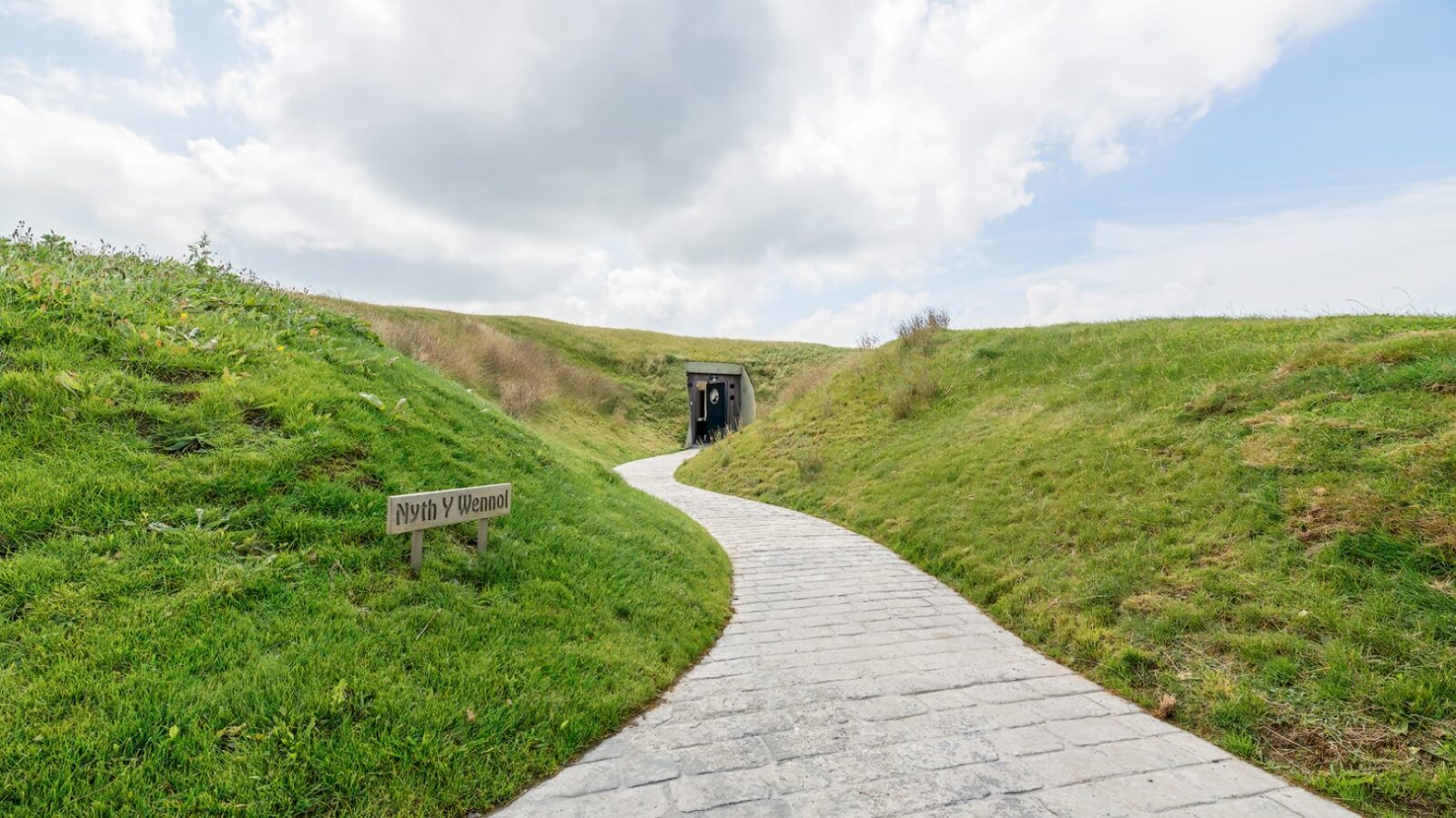 A curving stone path leads to The Willow Hill bunker entrance, set into grassy mounds under a partly cloudy sky. A sign on the left reads 