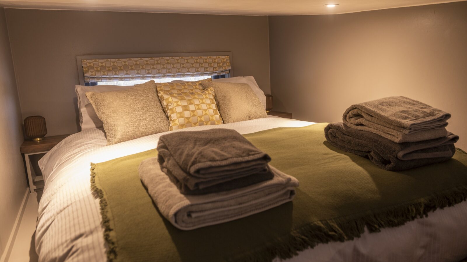 A cozy bedroom in a tiny house features a neatly made double bed, adorned with a white and green throw blanket, and several pillows. Two folded towel sets are placed on the bed. Soft, warm lighting illuminates the room, which has a small window with a patterned curtain behind the bed.