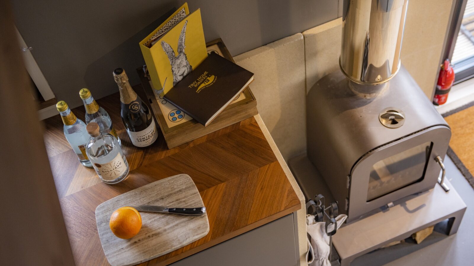 A cozy kitchen scene in a tiny home shows a wooden countertop with a cutting board holding a halved orange and a knife. Beside it are bottles of beverages and books. A metallic wood-burning stove to the right radiates warmth, making the space feel welcoming despite its borders.