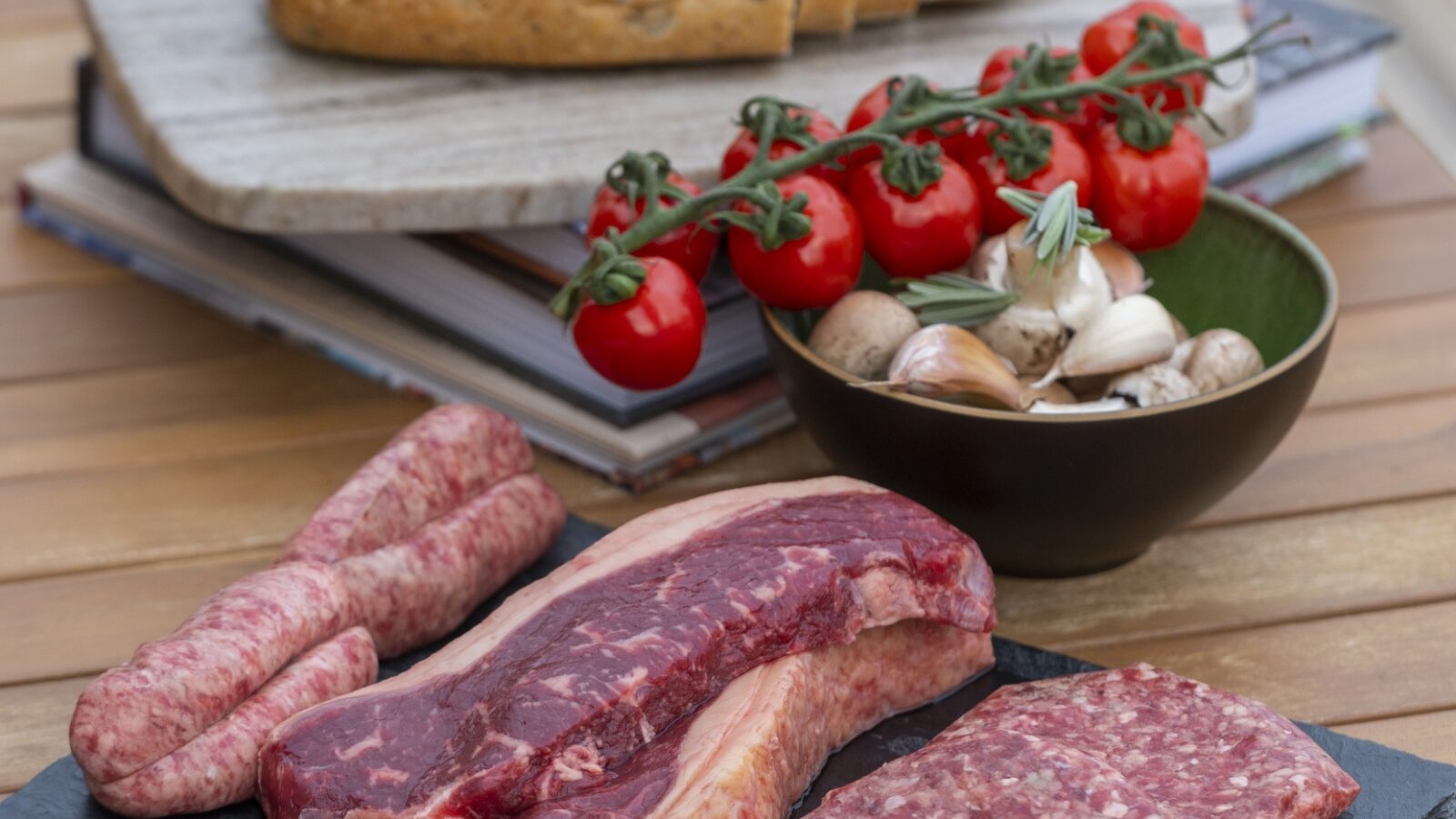 An assortment of raw meats, including sausages, steak, and burger patties, is displayed on a black slate on a wooden table. Next to it is a bowl of cherry tomatoes on the vine and mushrooms. A loaf of sliced bread sits on a cutting board in the background—a perfect setup for any tiny home kitchen.