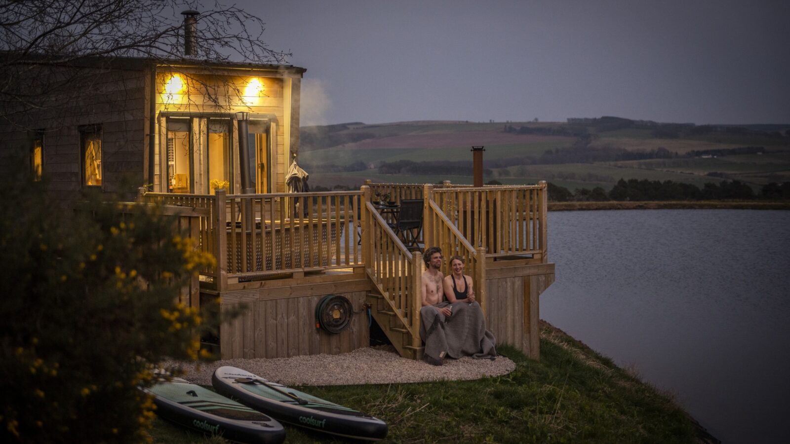 A couple sits wrapped in blankets on the steps of a cozy wooden cabin with illuminated windows, bordering a serene lake at dusk. The smoking chimney adds warmth, and two paddleboards rest nearby. Rolling hills frame this idyllic tiny home scene in the background.