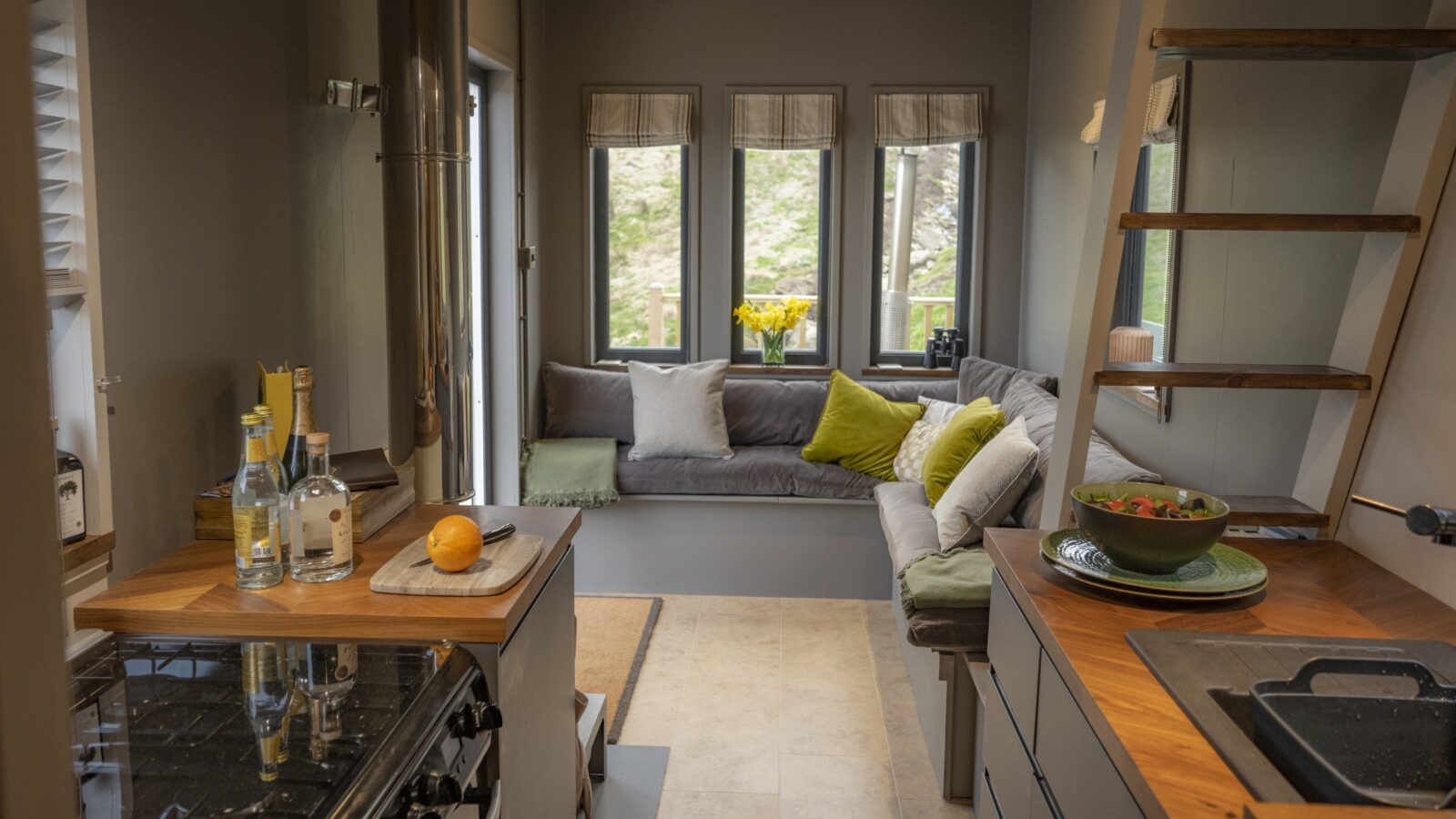 A modern tiny home interior featuring a compact kitchen with a gas stove and wooden countertops on the left. The living area at the back has a cozy built-in bench with green and yellow cushions, and windows with views of greenery. A ladder leads to a loft area, seamlessly blending borders of function and style.