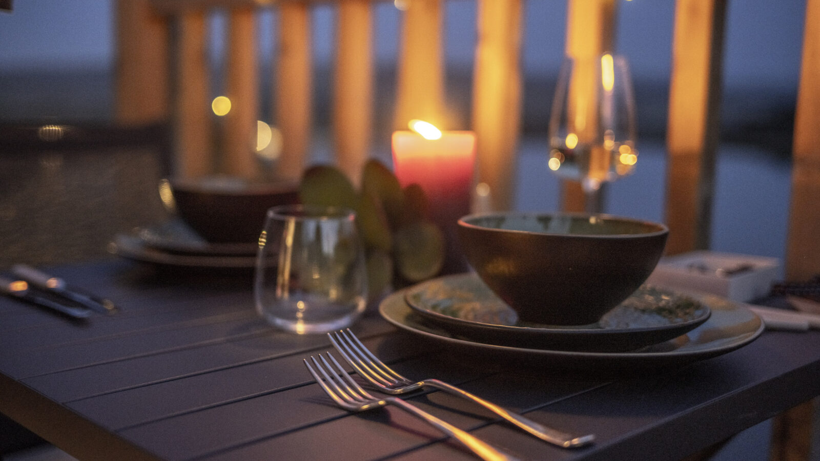 A cozy candlelit dinner setup on a tiny home's patio features a table with bowls, plates, glasses, and utensils. Soft string lights adorn the wooden railing in the background, creating a warm and intimate atmosphere that knows no borders during the evening.