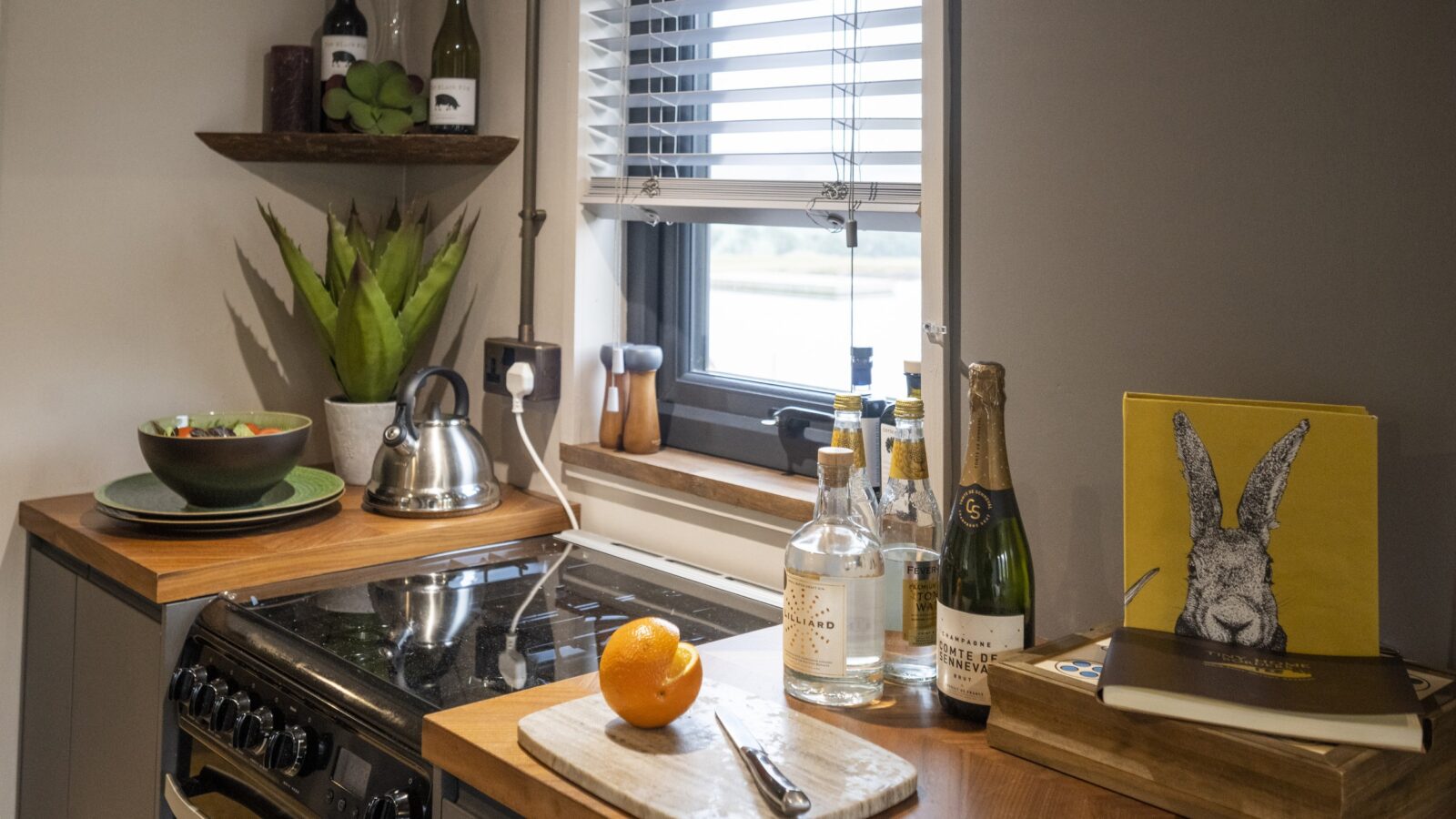 A cozy kitchen with a wooden countertop features a stove, a kettle, and assorted bottled drinks next to a window with blinds. In this tiny home, you'll find a cutting board with an orange, a knife, and a rabbit-themed cookbook. Shelves border the space with plants and wine bottles.