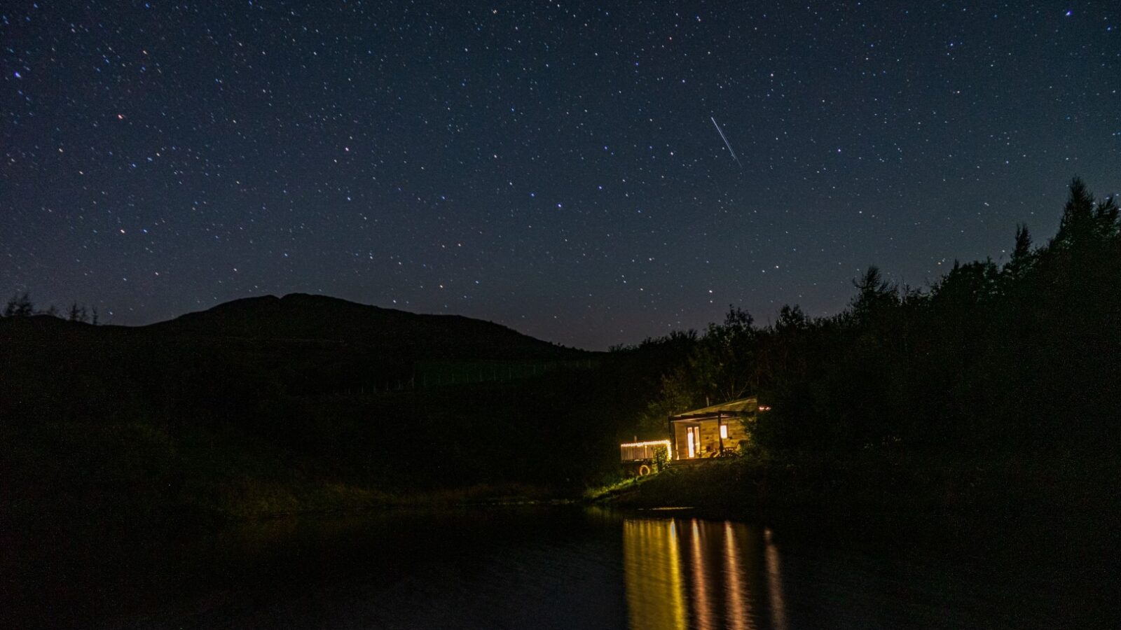 A tiny home with illuminated windows sits next to a calm lake under a star-filled night sky. The reflection of the home's lights shimmers on the water. A hill is silhouetted against the sky in the background, enhancing the serene and tranquil atmosphere within these borders.