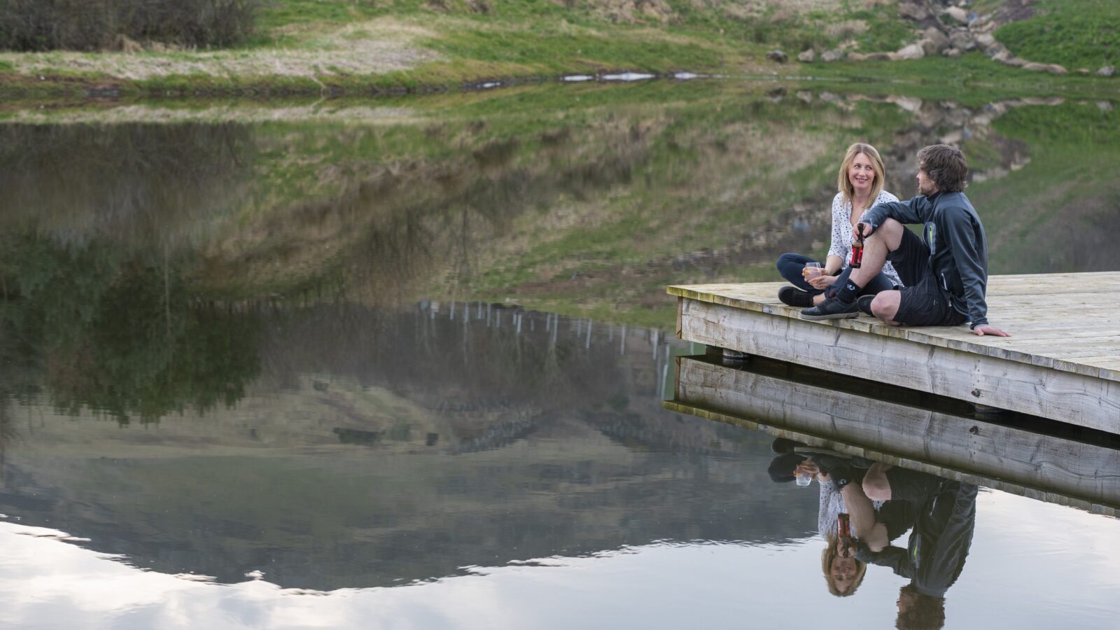 Two people sit on a wooden dock, their legs hanging over the edge, chatting and smiling. The dock extends into a calm lake, perfectly reflecting the hillside, greenery, and the individuals. Nearby, a quaint tiny home borders the lush green hills, enhancing the serene atmosphere.