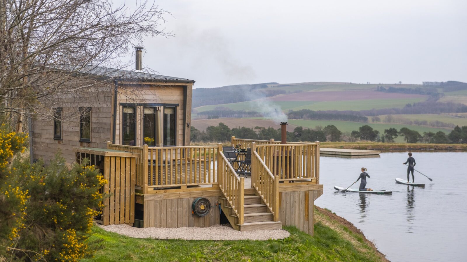 A wooden cabin with a deck sits beside a calm lake. Smoke rises from a chimney. Two people in wetsuits are paddleboarding on the lake. A grassy field and hills are visible in the background, framing this serene and natural scene of tiny homes nestled by nature's beauty.