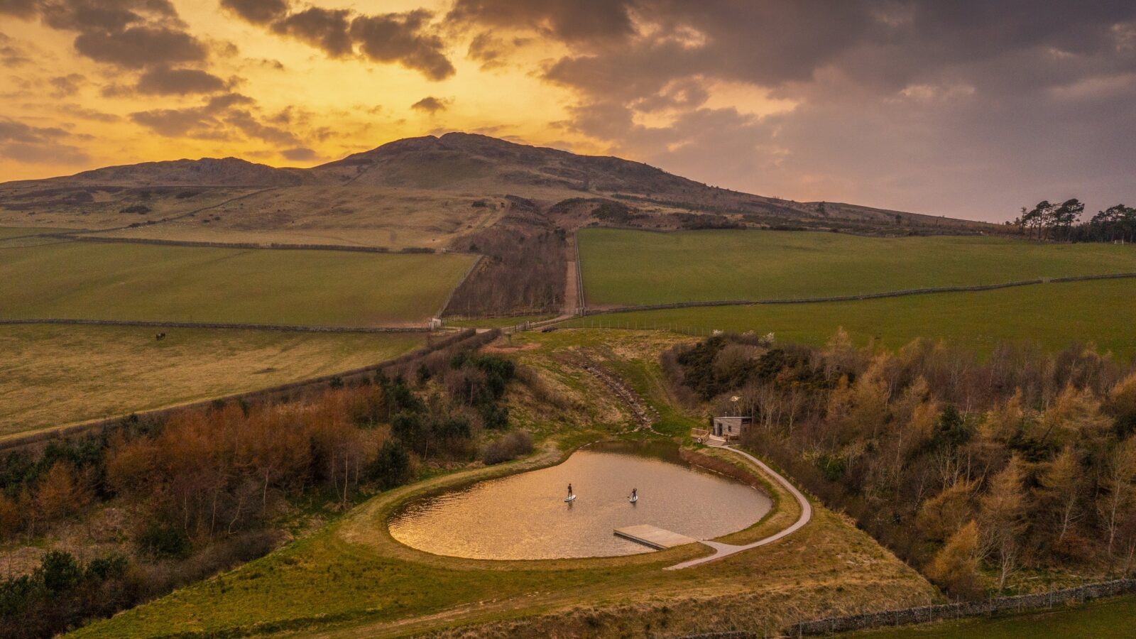 A small, heart-shaped pond with two fountains is surrounded by green fields and a few scattered trees, creating serene borders for a cozy tiny home nearby. A scenic hill stands in the background under a dramatic, colorful sunset sky with clouds and patches of golden light. A dirt path leads towards the pond.