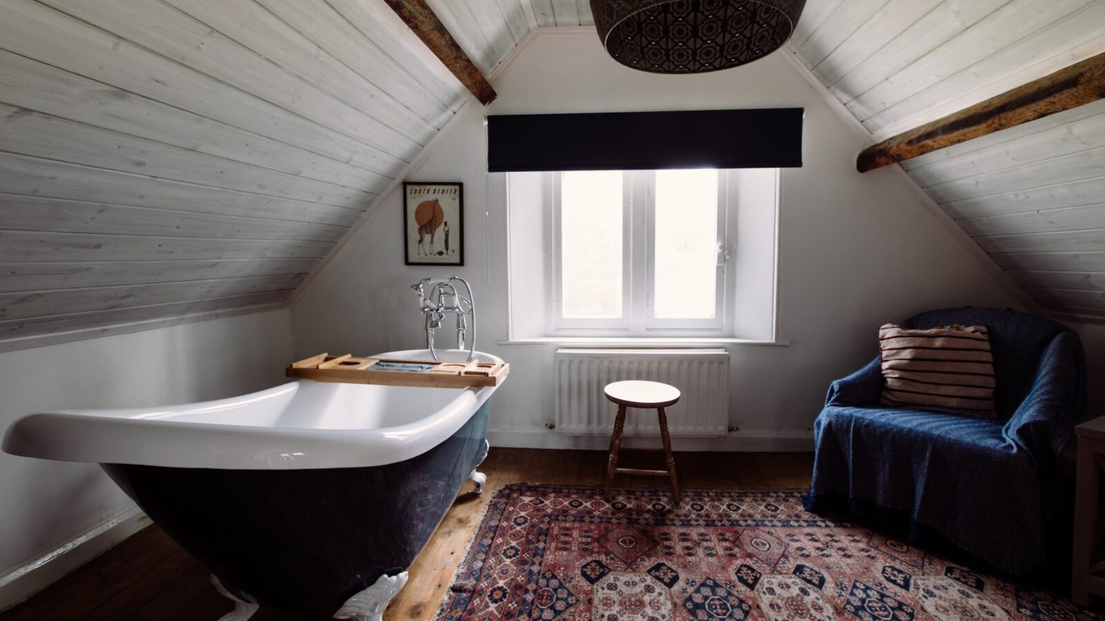 A cozy Top Lodge attic bathroom with white shiplap walls and a wooden floor features a freestanding black clawfoot bathtub with a wooden tray, a small wooden stool, a blue armchair with a striped pillow, a patterned rug, a ceiling light fixture, and a large window.