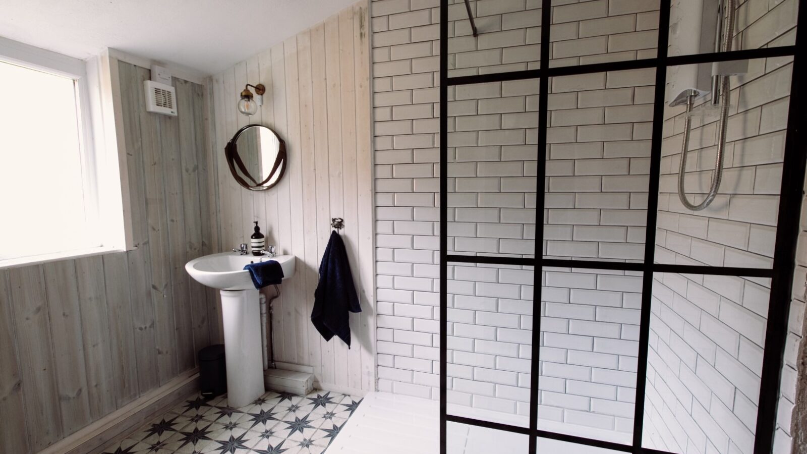 A modern bathroom at Top Lodge features light wood-paneled walls, a white sink with a round mirror above it, and a black towel hanging nearby. The shower area boasts a clear glass door with black trim and white subway tile walls. The floor is tiled with a striking black and white pattern.