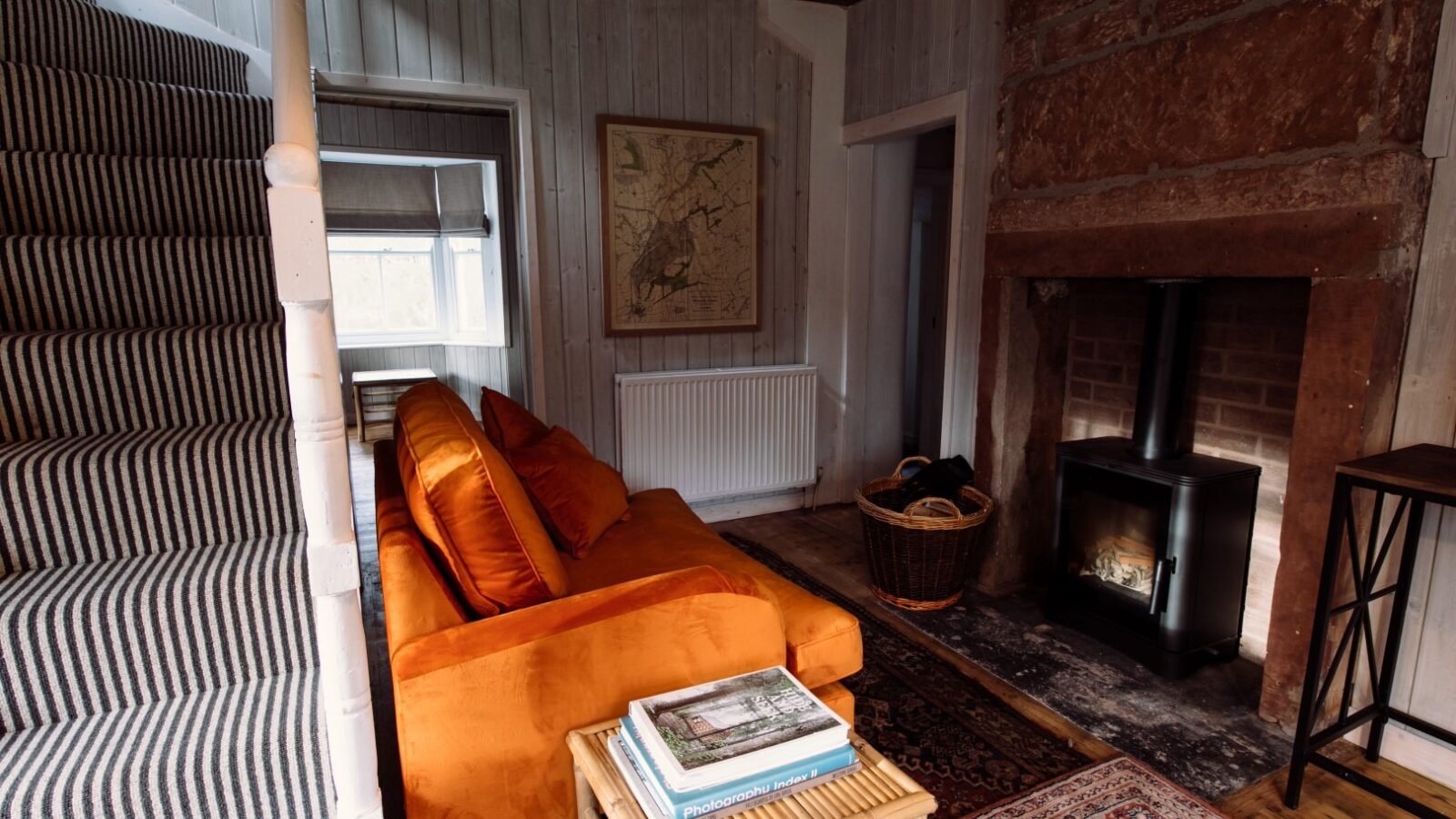 Cozy living room with a burnt orange sofa, a striped staircase to the left, a wood-burning stove on the right, and a map framed on the back wall. A wicker basket and side table with books are next to the stove. The floor, topped with a patterned rug, adds warmth to this lodge-like retreat.