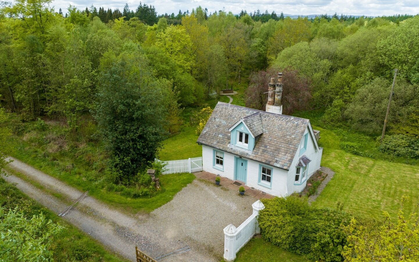 An aerial view of Top Lodge reveals a quaint cottage with a light blue exterior, nestled in lush greenery. The cottage boasts a slate roof, two chimneys, and a small white picket fence. A gravel driveway leads to the entrance, surrounded by trees and a neatly trimmed lawn.