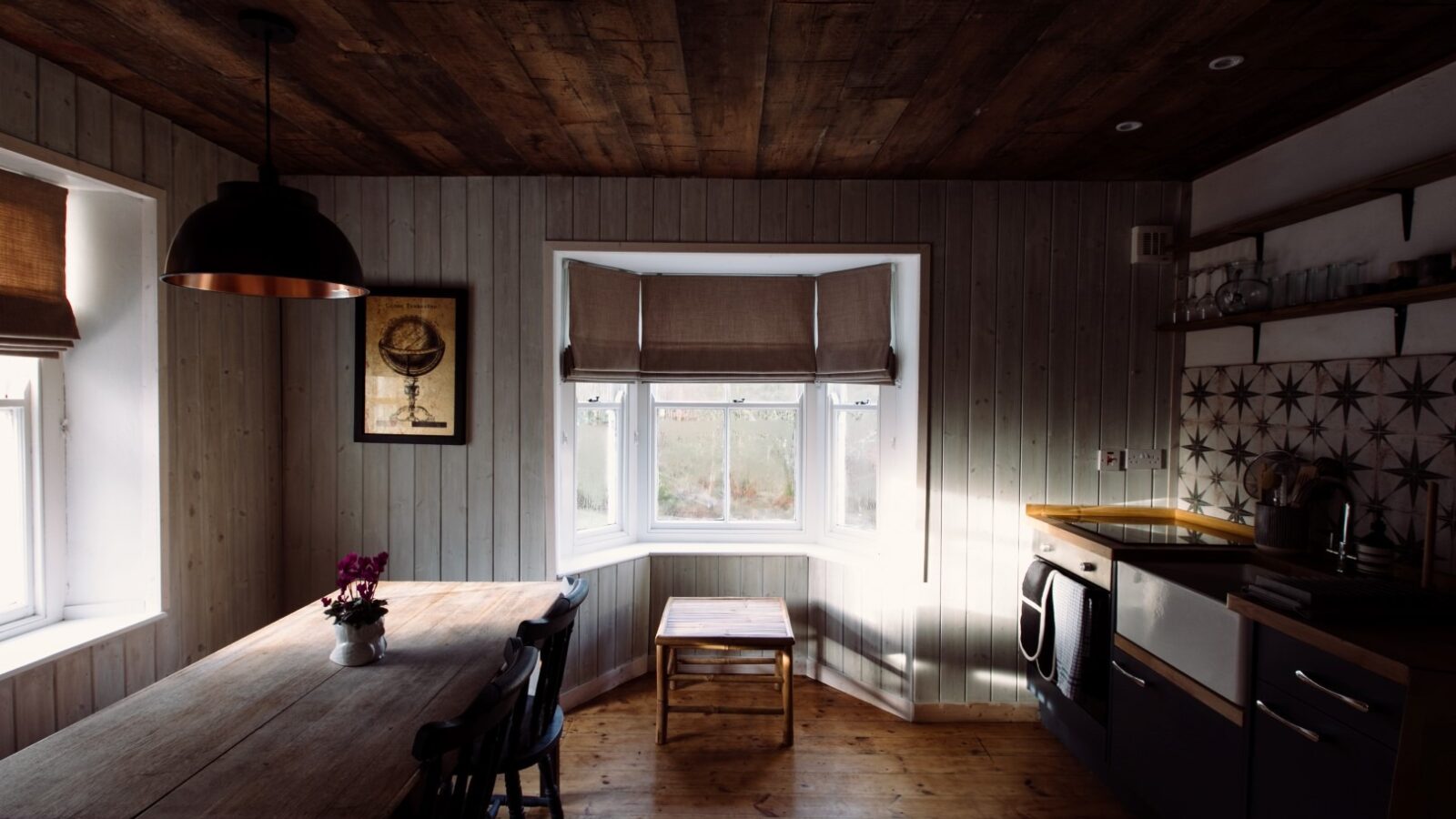 A cozy, rustic kitchen at Top Lodge features wooden walls and ceiling, illuminated by natural light from a bay window with fabric shades. The space includes a wooden dining table with chairs, a stove, a sink, and shelves with dishes. A framed artwork hangs on the left wall.