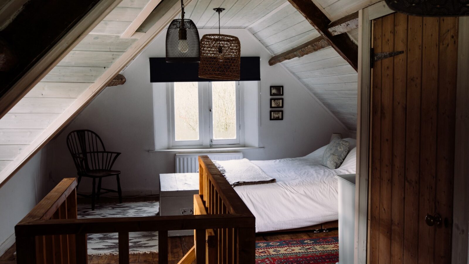 A cozy attic bedroom at Top Lodge with white-paneled walls and ceiling, featuring a bed with white sheets, a wooden chair, a bedside table with a lamp, and a colorful rug. The room has a window with a dark blind, wooden railings, and two pendant lights hanging from the ceiling.