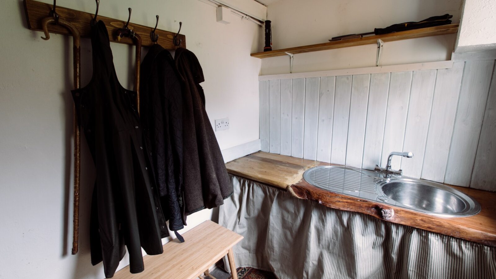 A cozy utility room with a wooden shelf holding various items, hooks with hanging coats and aprons, and a rustic counter with a stainless steel sink. The counter's natural wood edge adds charm, while the area underneath is hidden by a striped curtain. This top-notch design brings lodge-like warmth to the space.