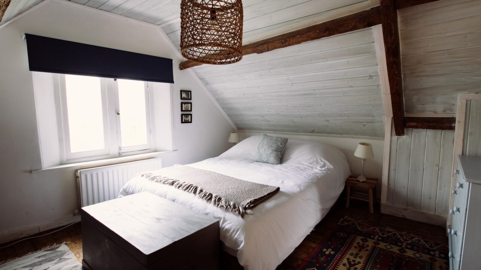 A cozy attic bedroom at Top Lodge with white wooden walls and a slanted ceiling. The room features a queen-sized bed topped with a beige throw blanket and a blue cushion. A wicker pendant light hangs from the ceiling. A wooden chest sits at the foot of the bed, and a colorful rug is on the floor.