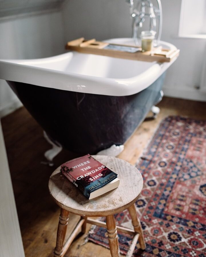 A cozy bathroom in Top Lodge style features a vintage black clawfoot bathtub on a wooden floor. A wooden stool in the foreground holds three books, with 