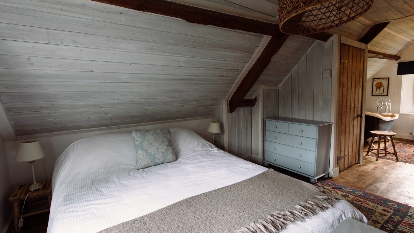 A cozy lodge attic bedroom with a slanted wooden ceiling and exposed beams. The room features a large bed with white linens and a patterned pillow, a gray dresser, two small lamps, and a wicker light fixture. An open door in the background leads to another room on the top floor.