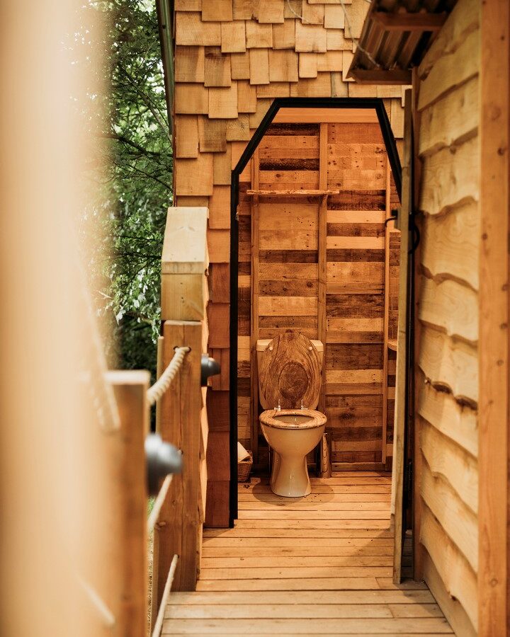 A narrow wooden hallway leads to a small rustic bathroom with a wooden door frame. The bathroom features a toilet with a wooden seat and backrest, surrounded by wood-paneled walls and a wood-shingled exterior. From Pennard Hill, you can see tree tents nestled among the trees in the background.