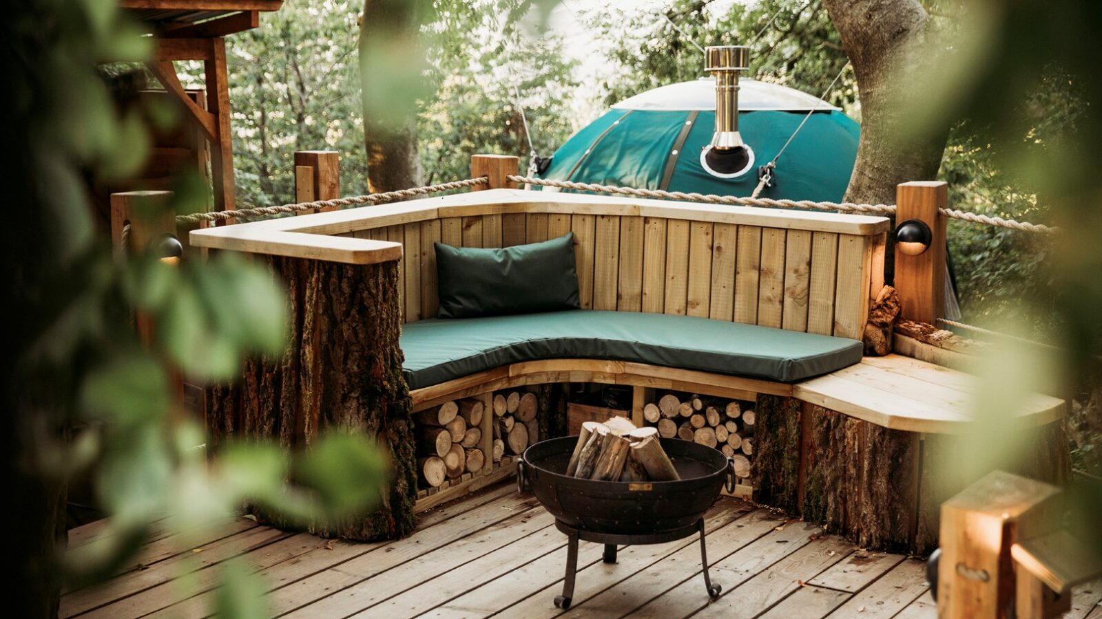 A cozy outdoor seating area nestled among trees at Pennard Hill features a corner bench with green cushions, crafted from wood with tree trunk accents. In front of the bench is a round fire pit filled with logs. The setting, enhanced by nearby tree tents, conveys a rustic, inviting ambiance.