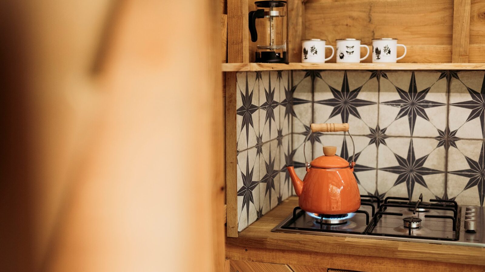 A cozy kitchen with wooden cabinets features a tiled backsplash with a star pattern. An orange teapot sits on a gas stove with a blue flame. A French press and three mugs with black designs are neatly arranged on a shelf above the stove, evoking the charm of Pennard Hill retreat spaces.