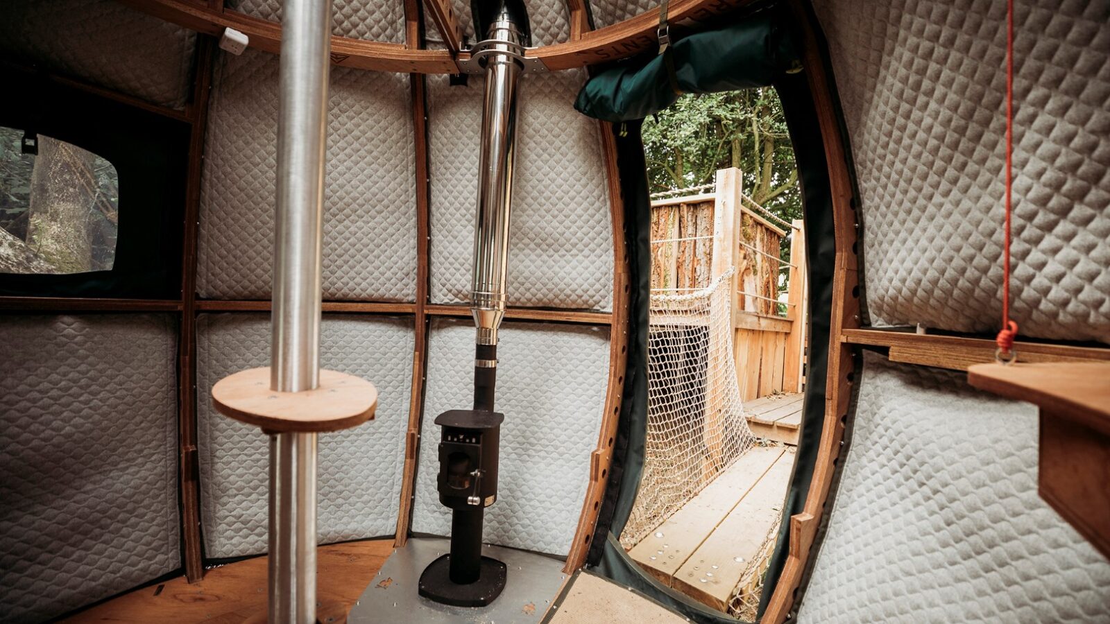 Interior view of a cozy yurt at Pennard Hill with quilted fabric lining the rounded walls, a central metal stove pipe extending from a small wood-burning stove, and a round doorway leading to an outdoor wooden deck. The space appears warm and rustic with natural light shining in.