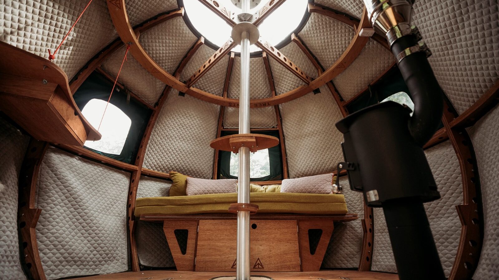 The image shows the interior of a cozy, dome-shaped wooden cabin nestled at Pennard Hill. It includes a cushioned bench, circular windows, and padded walls with insulating material. A central pole and a wood-burning stove are prominent features, creating a warm and inviting atmosphere reminiscent of Tree Tents.