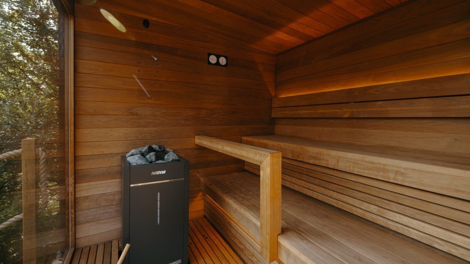 A wooden sauna room at Forcett Grange features a wooden bench along the right wall, a Harvia sauna heater with stones on the left, and a ladle and bucket on the floor. Soft lighting highlights the warm wood tones, creating a cozy and inviting atmosphere.