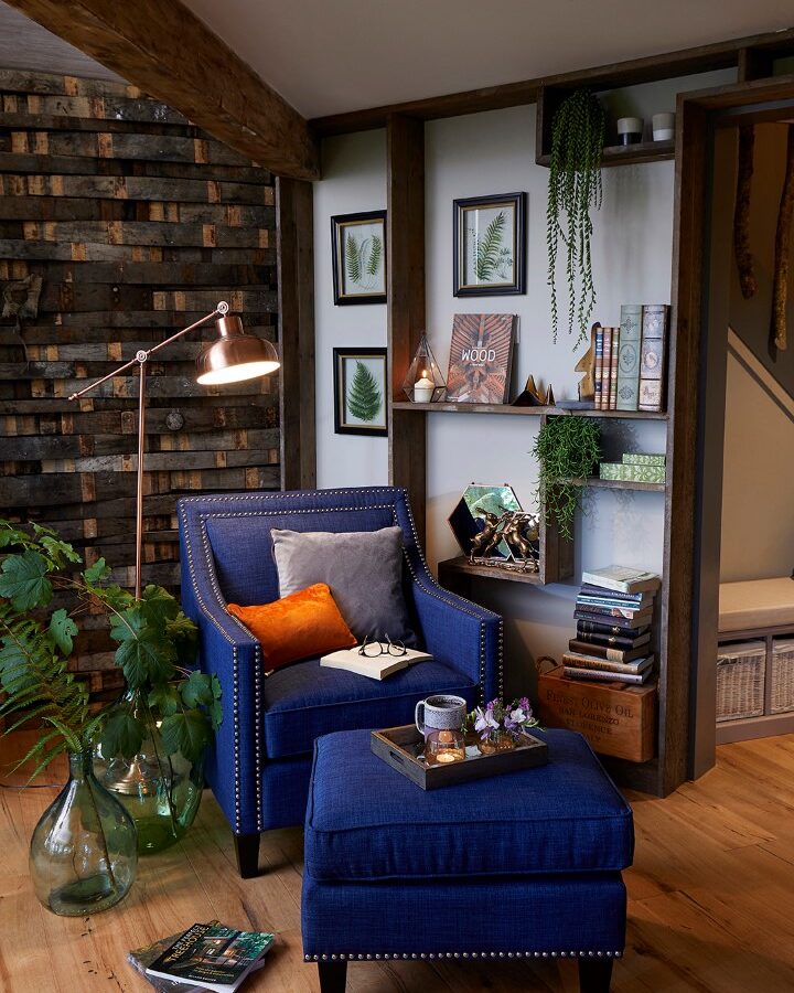 A cozy reading nook in the luxurious Ravendere Treehouse features a blue armchair with matching ottoman, complemented by wood accents and indoor plants. Adorned with gray and orange pillows, the chair sits next to shelves displaying books, a clock, and framed art. A floor lamp provides light.