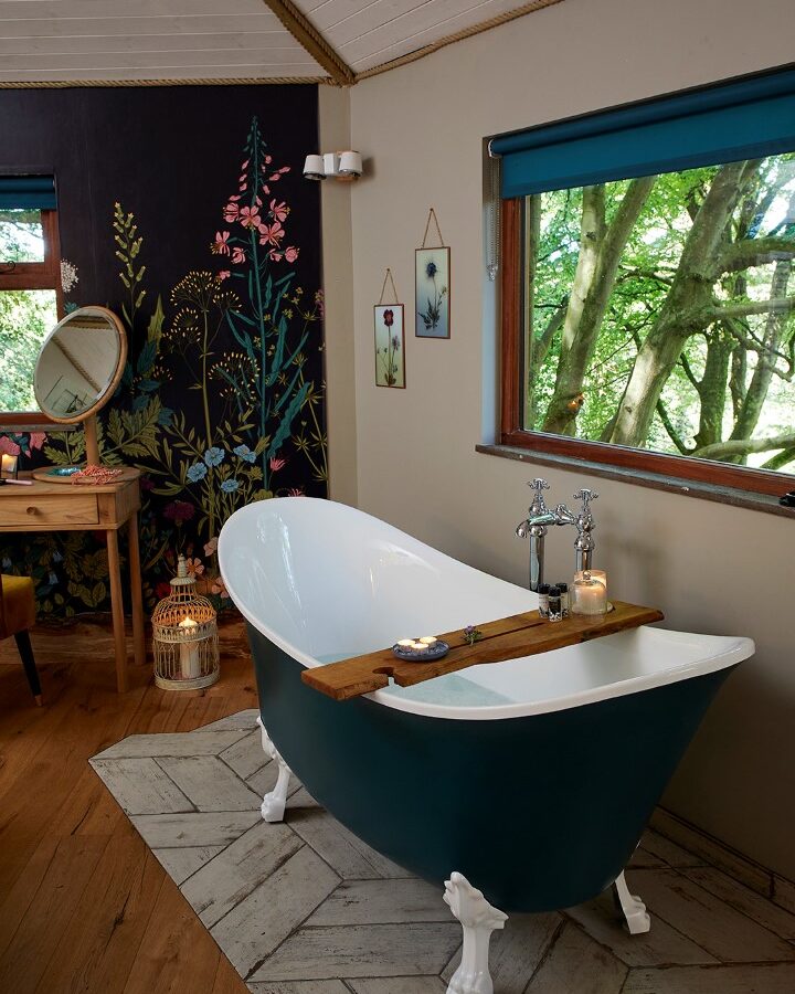 A cozy bathroom in Ravendere features a dark green clawfoot bathtub with a wooden bath tray, candles, and bath items. The room has a rustic wood floor, a vanity with a round mirror, and a mural with tall floral designs. Natural light streams through a large window showcasing treehouse-like views of green trees.