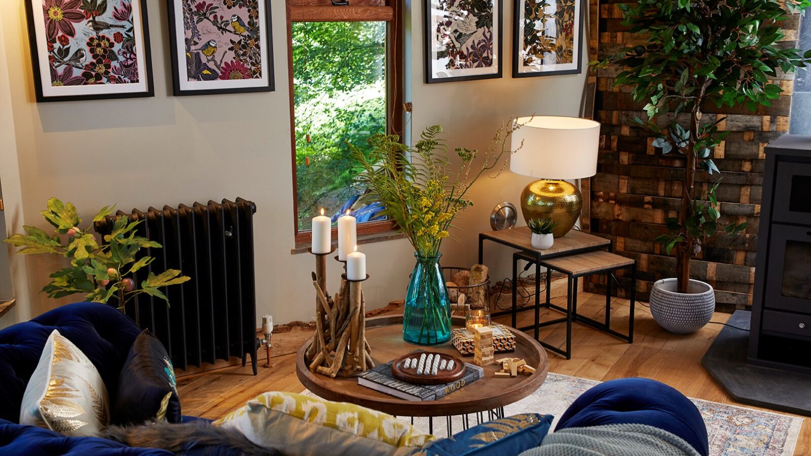 A cozy living room in the Ravendere Treehouse features a blue couch, a wooden coffee table with candles and decorations, a green vase with flowers, a modern lamp, and framed botanical art on the wall. To the right, a large plant and a wood-paneled accent wall complement the warm, natural decor.