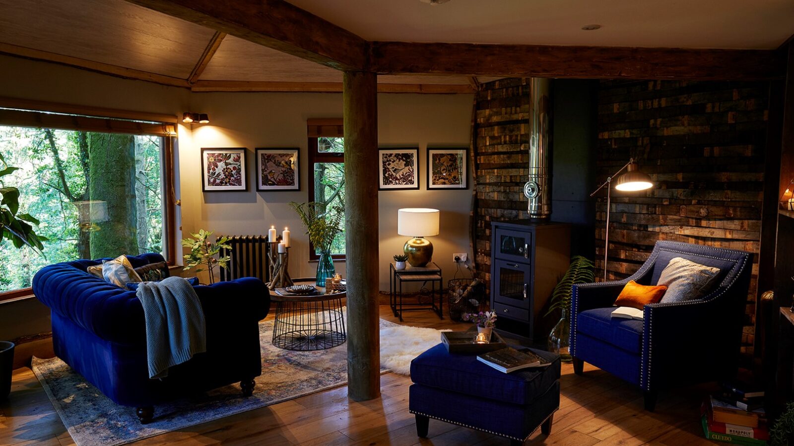 A cozy living room with a rustic feel, reminiscent of Ravendere Treehouse. The space features a blue velvet sofa and armchair, a wood-burning stove, wooden beams, a brick accent wall, and large windows with views of greenery. Warm lighting and various decorative items add to the inviting atmosphere.