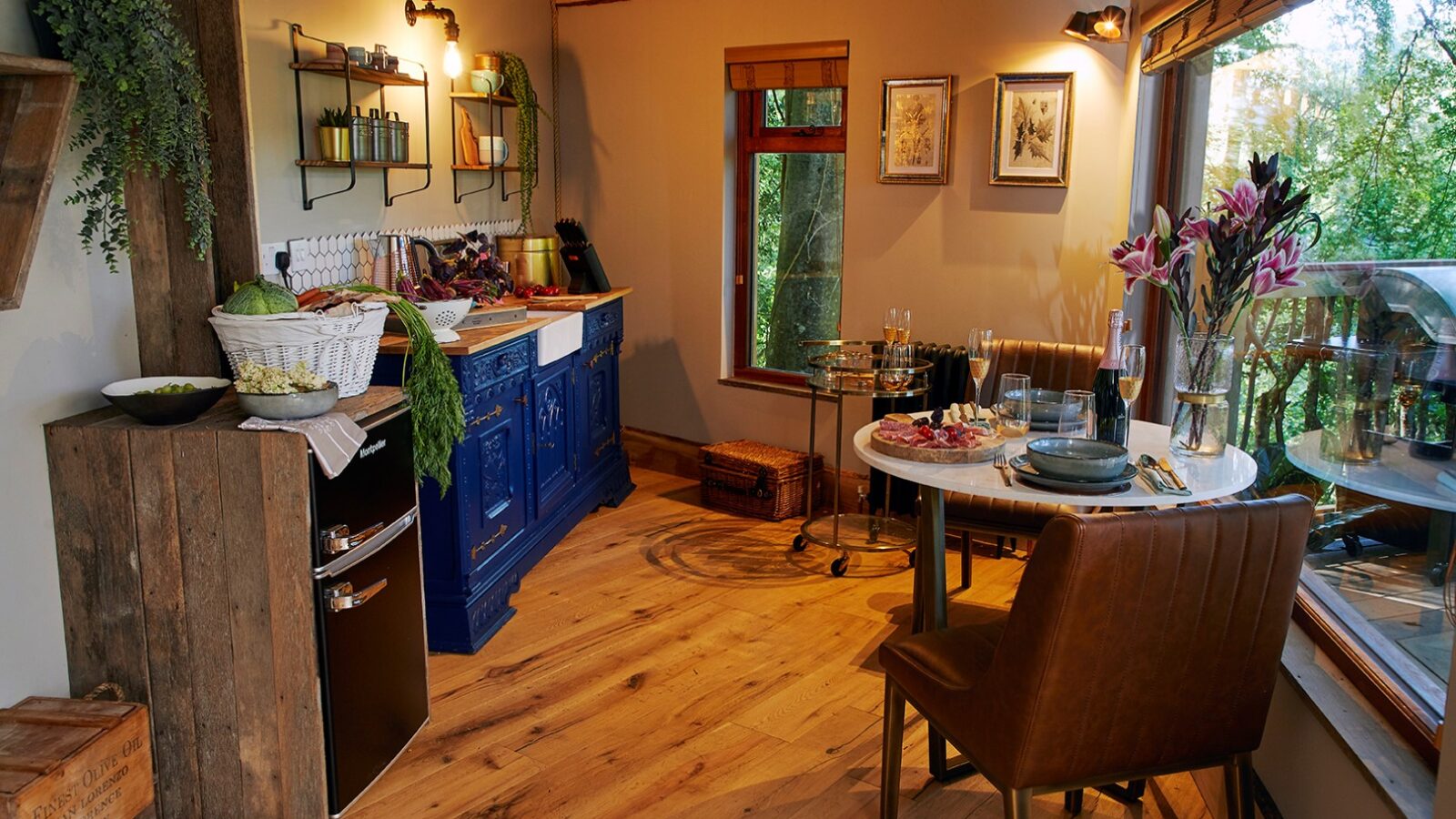 Cozy kitchen with wood floors and a blue stove in the charming Ravendere Treehouse. A small wooden table set with plates and wine glasses is next to a large window overlooking a lush garden. Shelves with plants and kitchen items hang on the wall, while a vase with lilies and framed pictures add decoration.