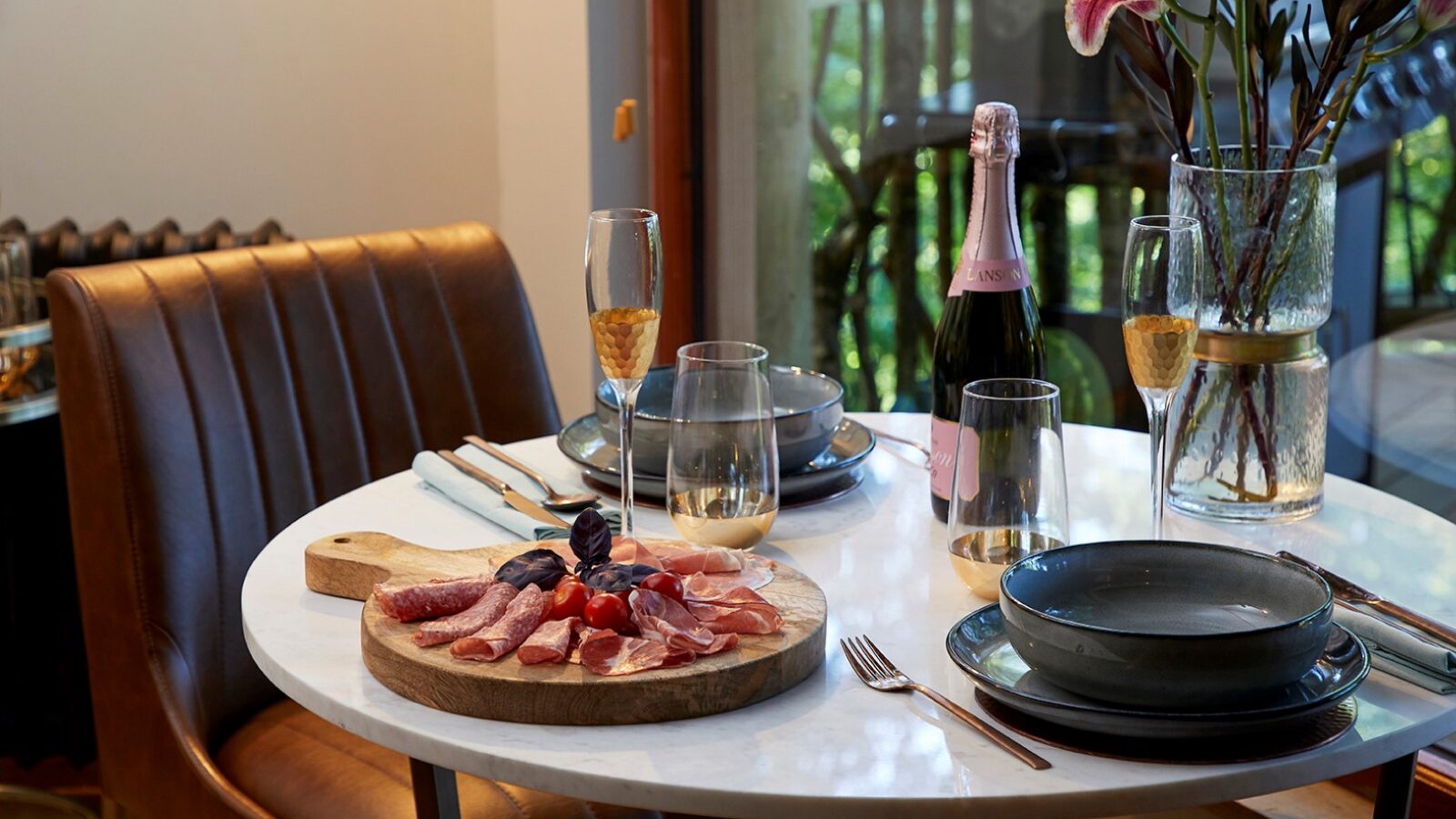 A cozy dining table at Ravendere Treehouse is set with a charcuterie board of meats, a bottle of pink champagne, two filled flutes, and elegant black dinnerware. The table is decorated with a vase of pink lilies, positioned near a sunny window with lush greenery visible outside.